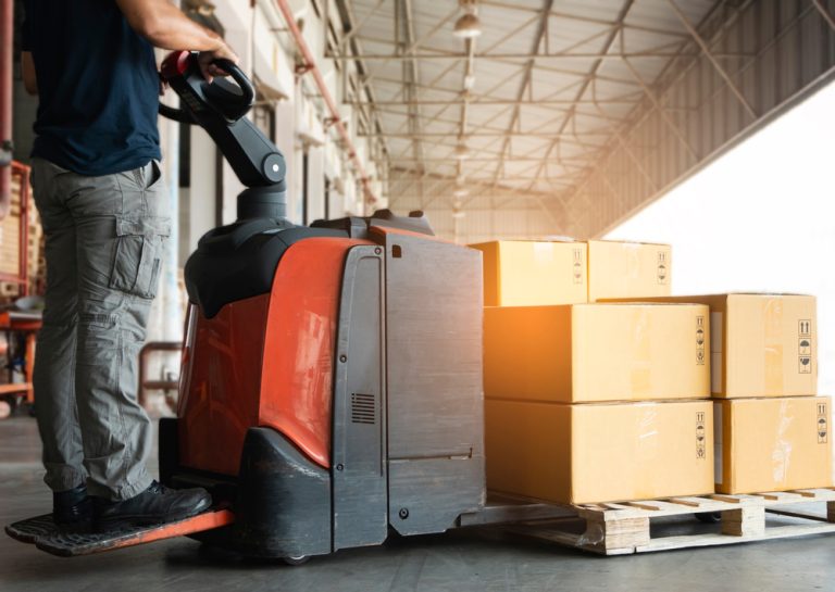 man standing on and operating electric pallet truck with boxes loaded on